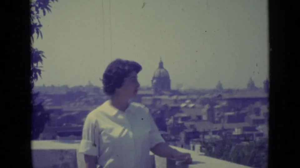 The woman appears in profile on a balcony above Vatican City, with rooftops and a prominent dome under a hazy sky.
