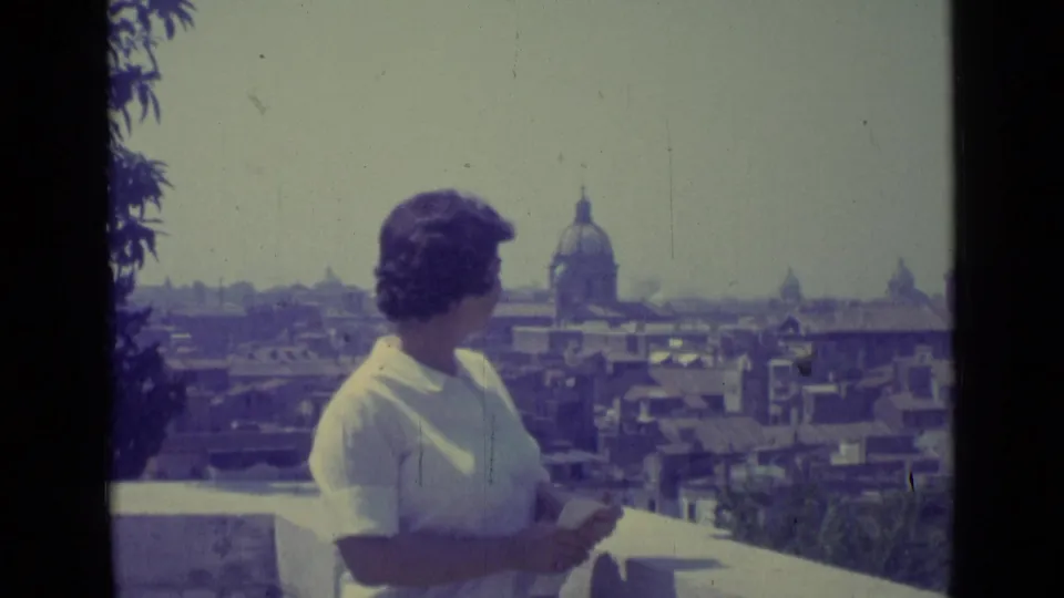 The woman faces the skyline from a balcony, with Vatican City rooftops and a domed landmark visible beyond her.