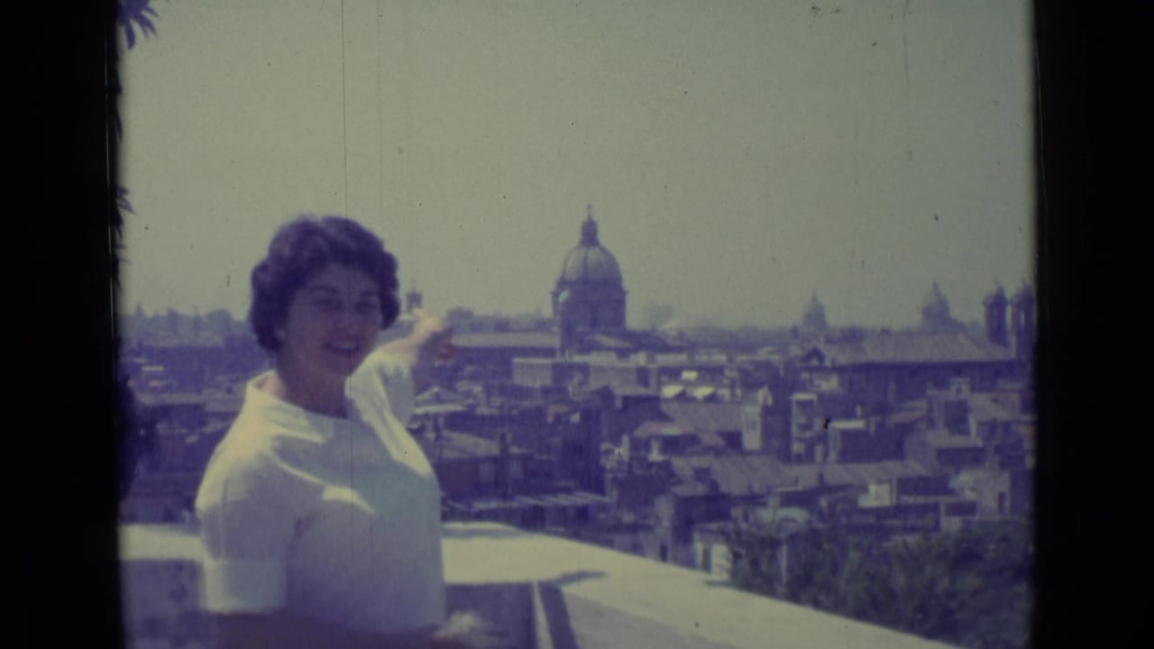 A smiling woman points from a balcony toward Vatican City rooftops and a prominent dome in the background.