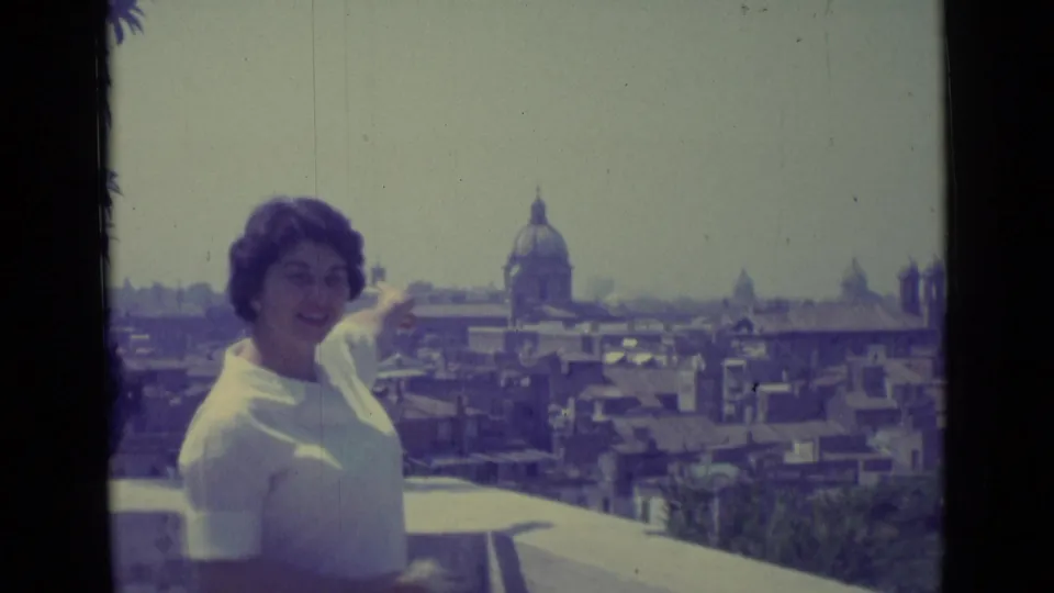 A smiling woman points from a balcony toward Vatican City rooftops and a prominent dome in the background.