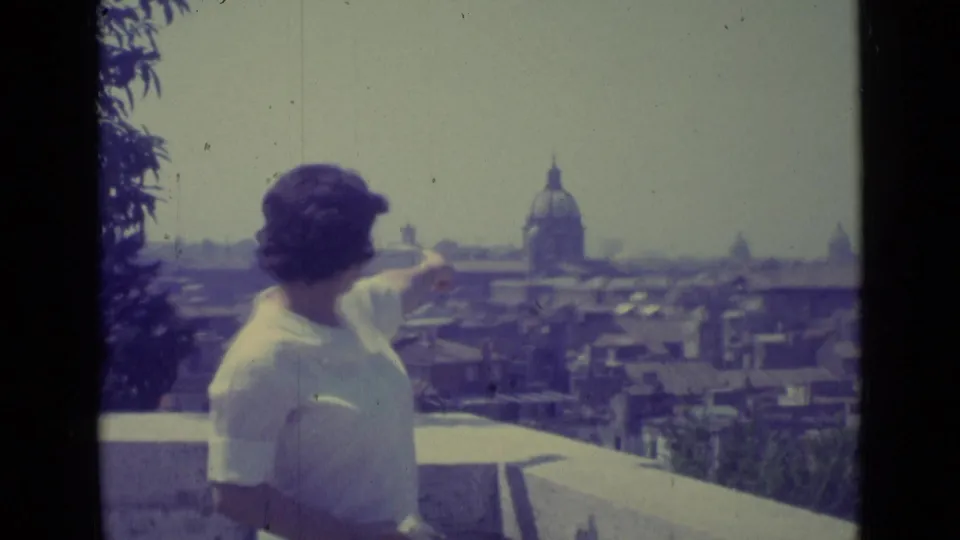 In 1962 Vatican City footage, a woman in a white blouse points across rooftops toward a prominent church dome from a terrace.