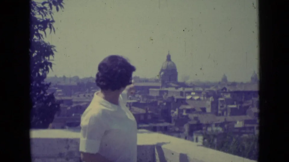 A woman in a white blouse stands at a Vatican City terrace as the domed church and surrounding rooftops remain in view.