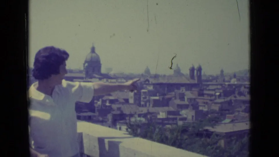 In 1962 Vatican City footage, a woman points across a balcony rail toward Rome rooftops and a large distant dome.