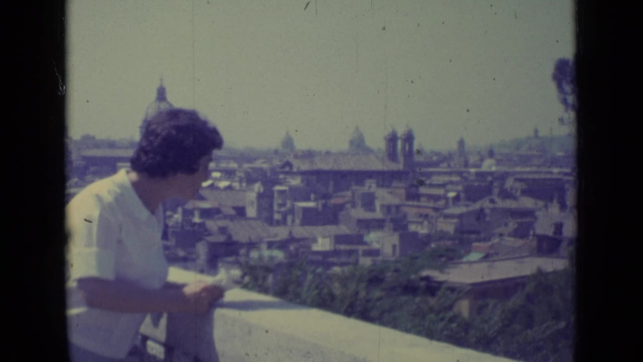 The woman stands at the balcony rail in 1962 Vatican City footage, looking over rooftops and distant domes.