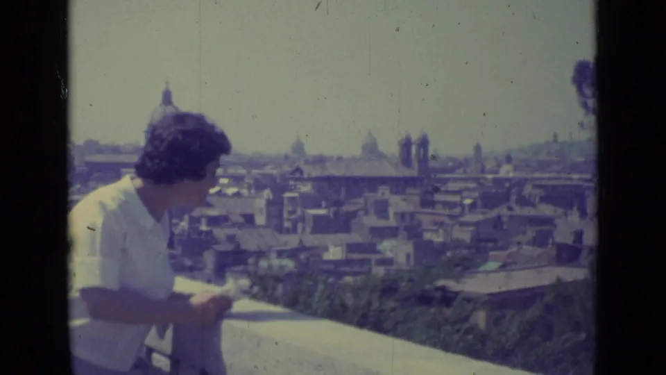 The woman stands at the balcony rail in 1962 Vatican City footage, looking over rooftops and distant domes.