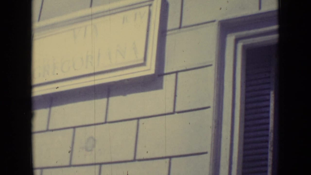 A close-up shows a pale masonry facade, a partly legible placard, and a dark shuttered window near Vatican City.