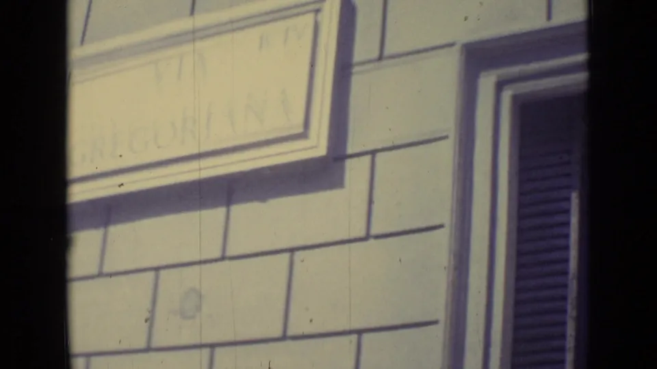 A close-up shows a pale masonry facade, a partly legible placard, and a dark shuttered window near Vatican City.