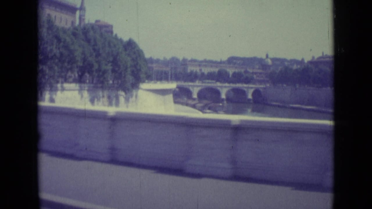 Moving 8mm view of a riverside road, tree line, and arched bridge in the Vatican City or Rome-area setting.