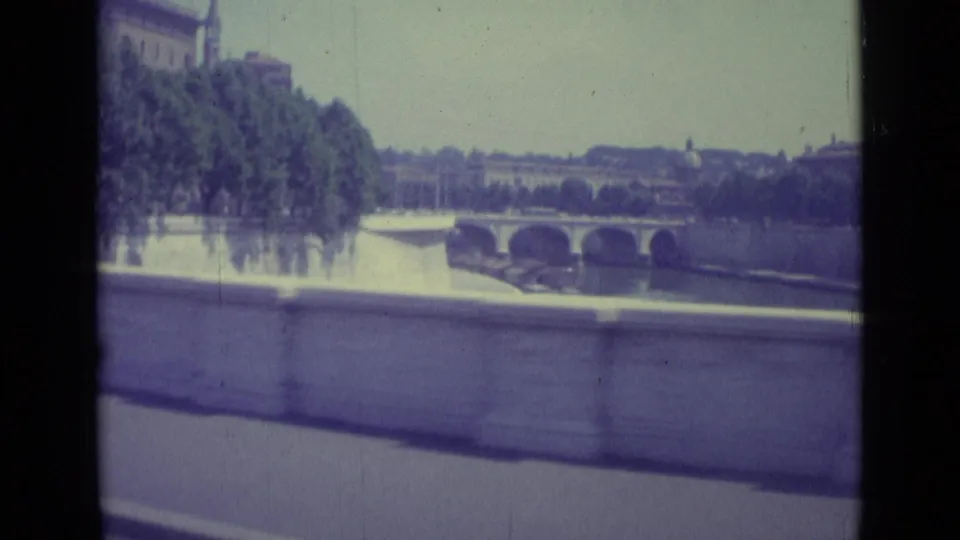 Moving 8mm view of a riverside road, tree line, and arched bridge in the Vatican City or Rome-area setting.