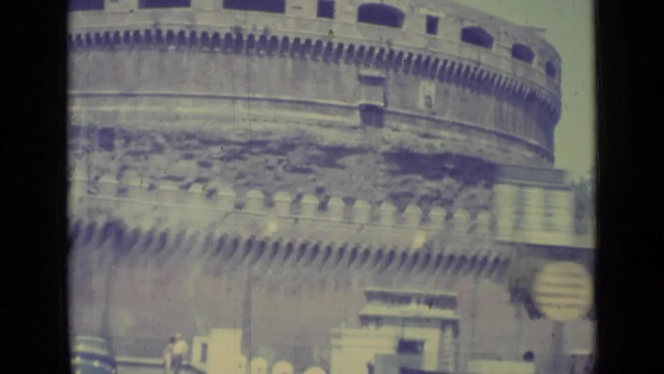 1962 8mm frame of Castel Sant'Angelo showing the cylindrical stone wall, battlements, street foreground, cars, and tiny visitors.