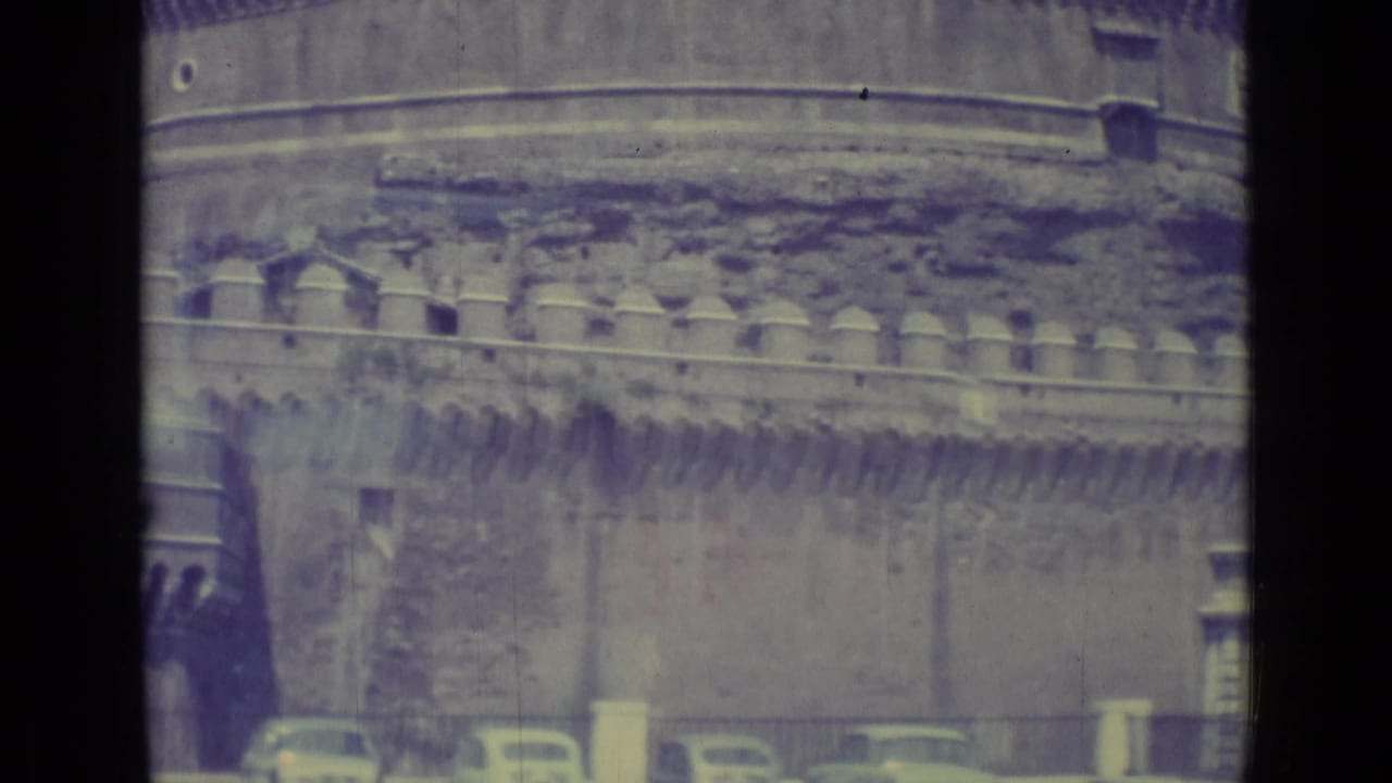 1962 8mm frame of Castel Sant'Angelo with crenellated stonework above a street lined with railings, trees, and parked cars.
