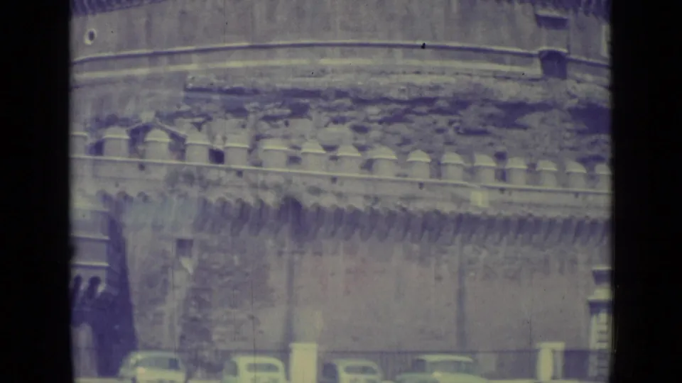 1962 8mm frame of Castel Sant'Angelo with crenellated stonework above a street lined with railings, trees, and parked cars.