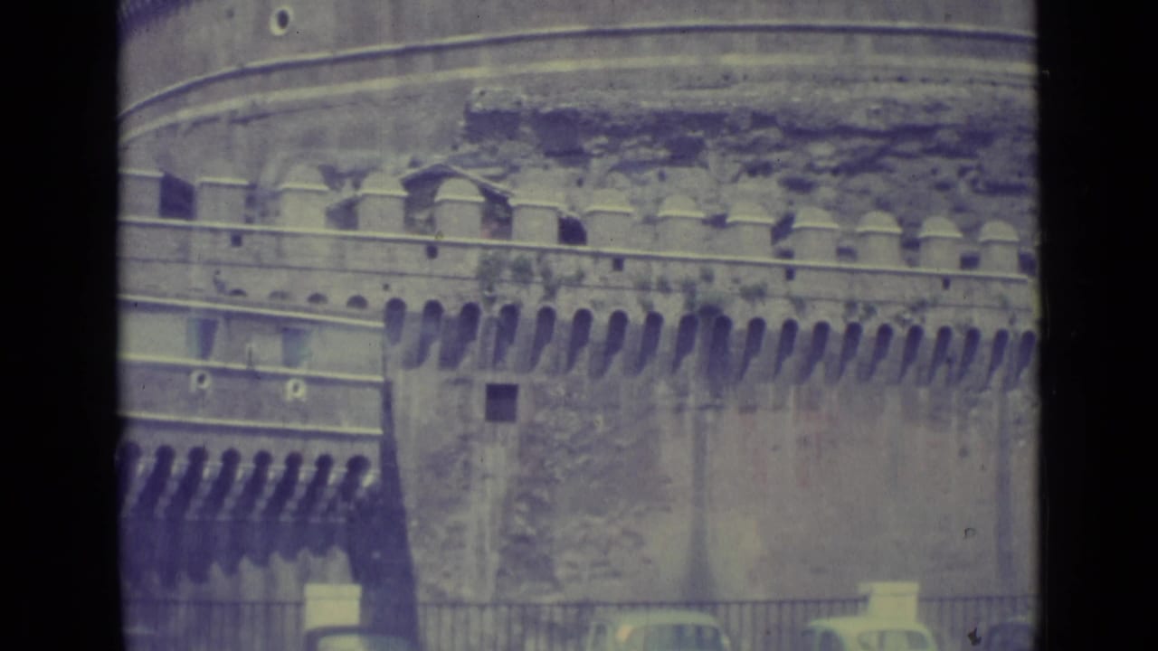 1962 8mm view of Castel Sant'Angelo's stone battlements and wall, with railings, trees, and cars partly visible at street level.