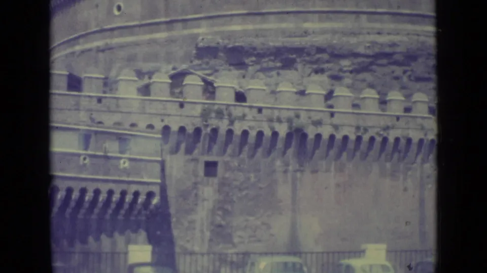 1962 8mm view of Castel Sant'Angelo's stone battlements and wall, with railings, trees, and cars partly visible at street level.