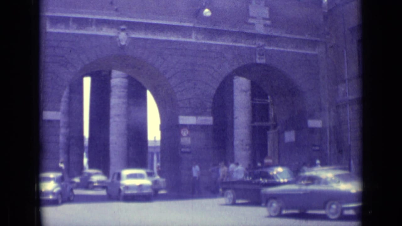 1962 Vatican City 8mm frame showing cars passing near monumental stone arches with pedestrians at the entrance.