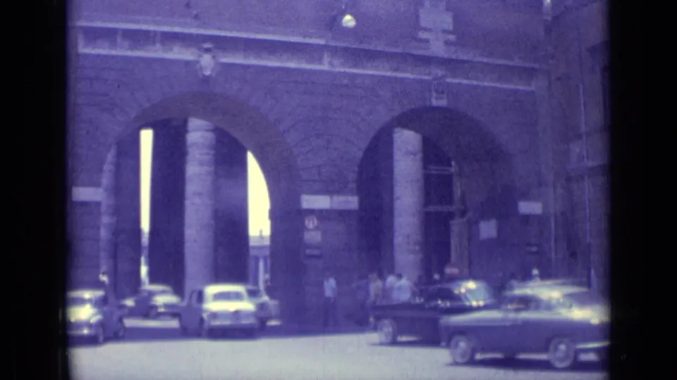 1962 Vatican City 8mm frame showing cars passing near monumental stone arches with pedestrians at the entrance.
