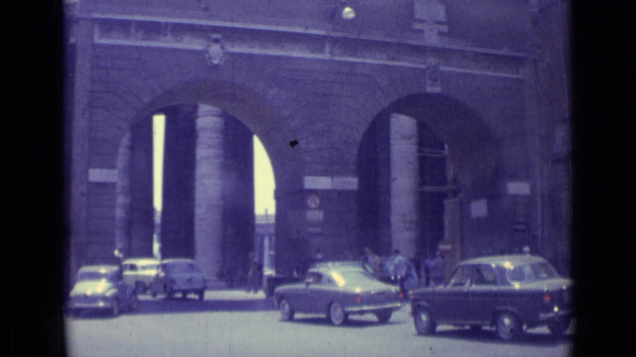 Several cars pass in front of tall arched openings with pedestrians nearby.