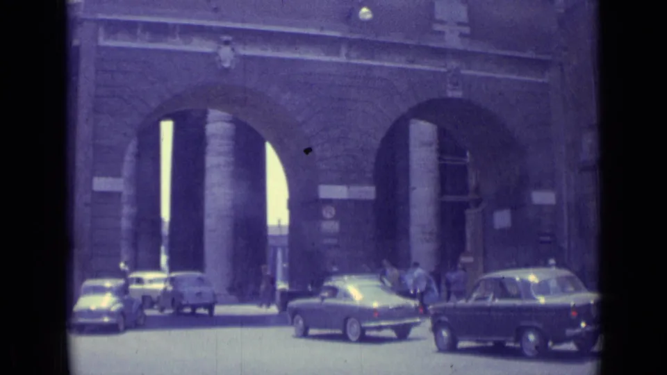Vatican City street view from 1962 with mid-century cars crossing before tall masonry arches and people near the entrance.
