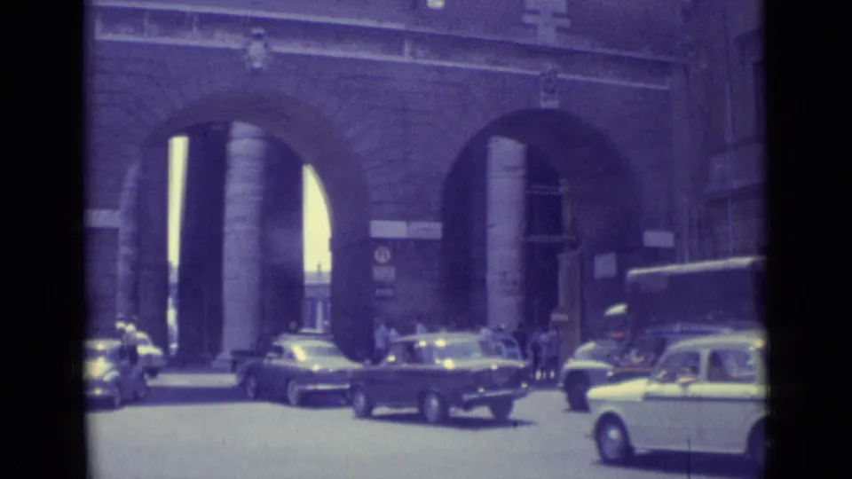 1962 Vatican City frame showing cars and a larger vehicle moving outside monumental arches as pedestrians gather near the passage.