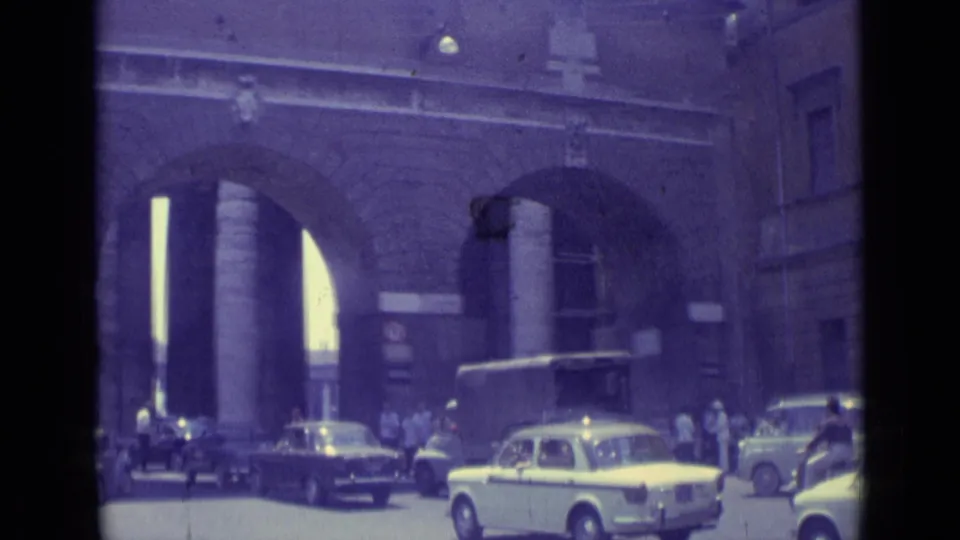 Vatican City 1962 home-movie frame with cars, pedestrians, and a bus-like vehicle near a monumental stone arch entrance.