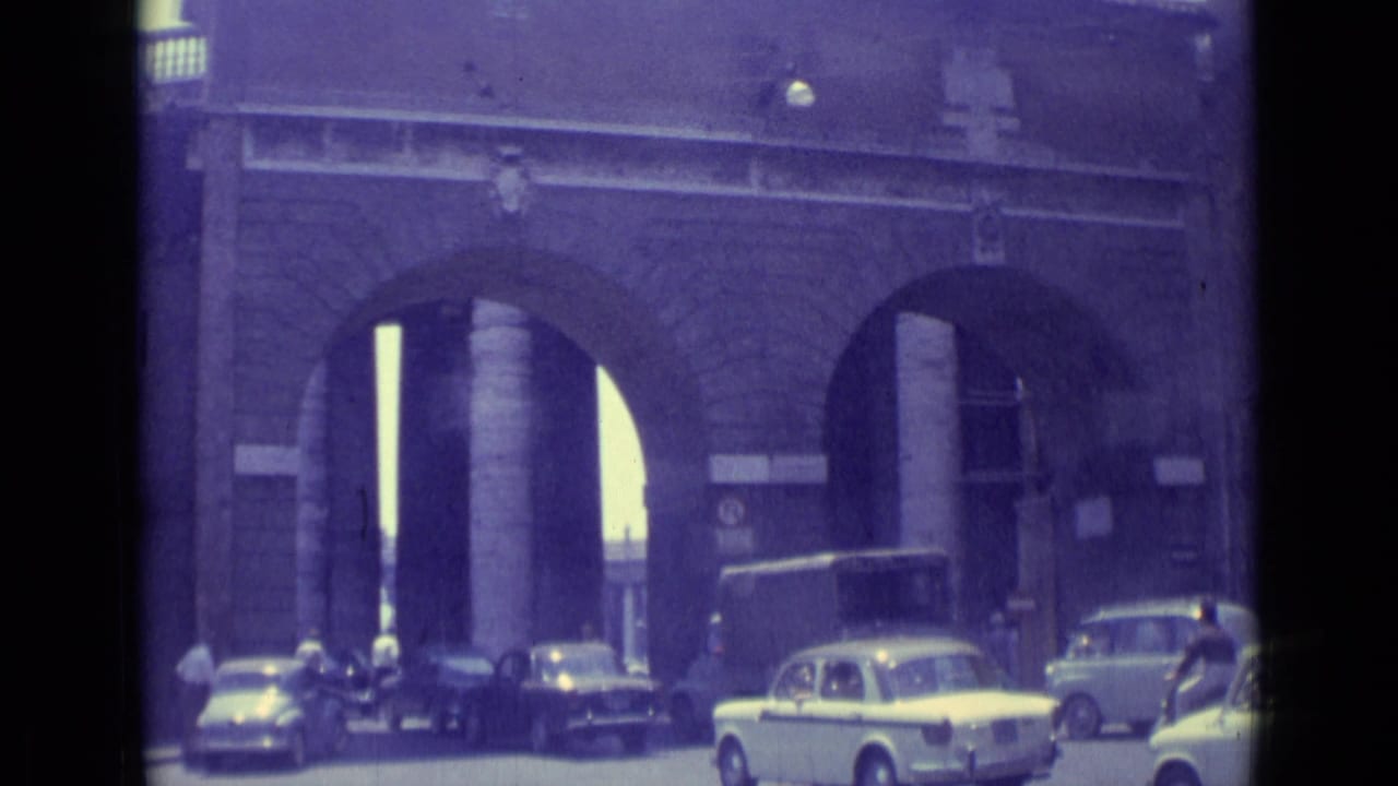 1962 Vatican City street view with classic cars moving near large masonry archways and pedestrians at the roadside.