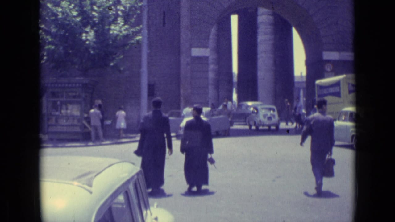 1962 Vatican City street scene with pedestrians in dark suits, parked and moving cars, trees, and a large arched masonry entrance.