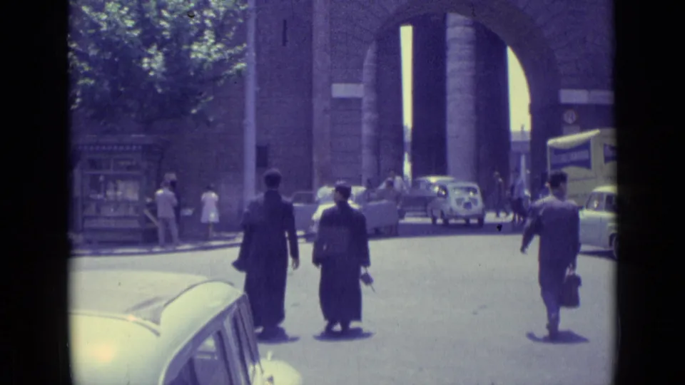 1962 Vatican City street scene with pedestrians in dark suits, parked and moving cars, trees, and a large arched masonry entrance.