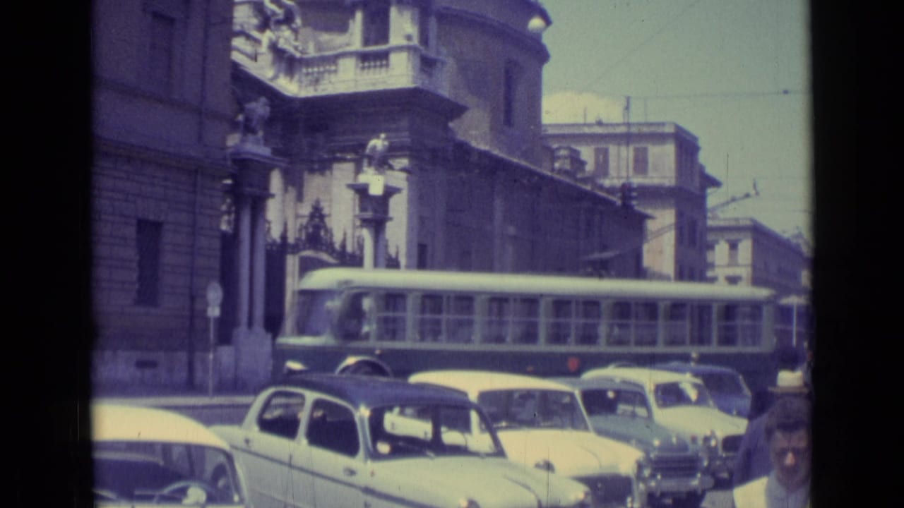 Long bus passing compact cars on a busy street beside ornate stone buildings.