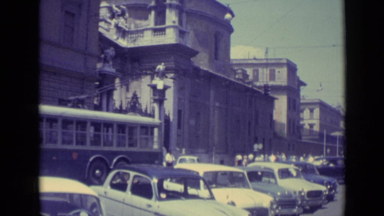 1962 Vatican City street view with the rear of a long bus, several compact cars, pedestrians, and a large stone building in shadow.