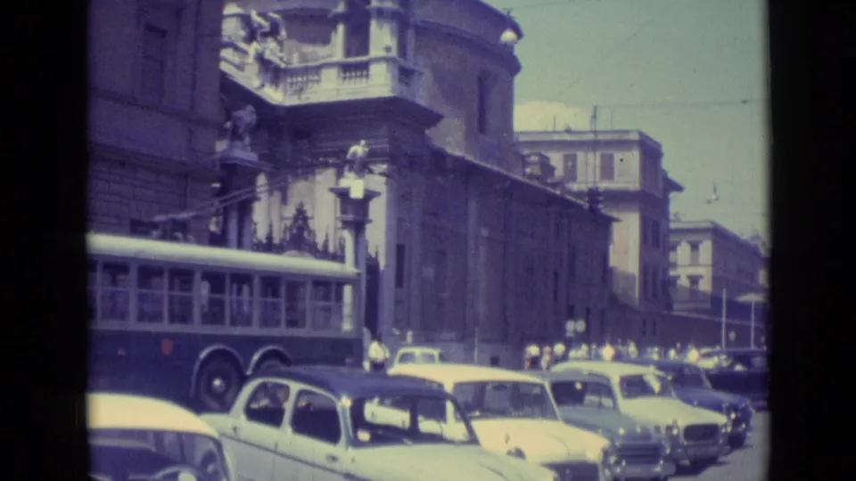 1962 Vatican City street view with the rear of a long bus, several compact cars, pedestrians, and a large stone building in shadow.