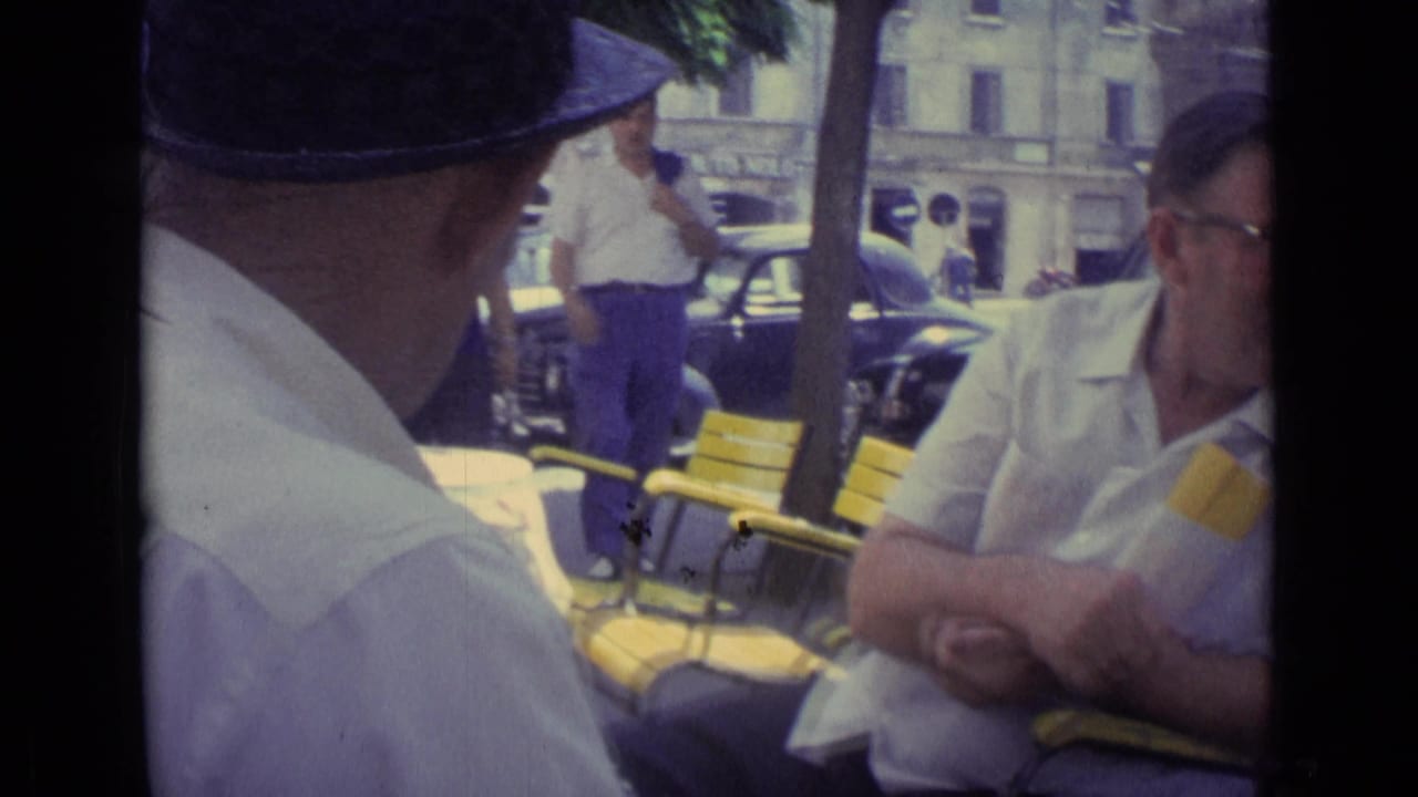 Two men seated at an outdoor table with yellow chairs and cars behind them.