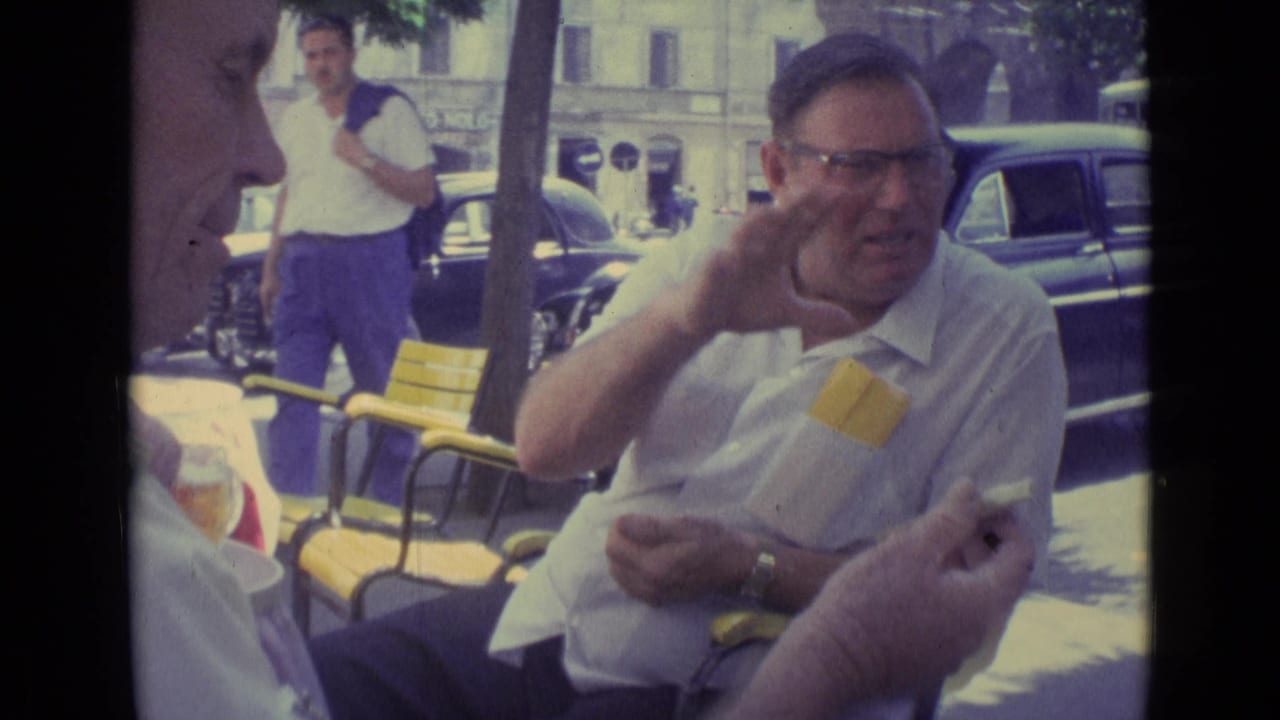 A man in glasses and a white shirt gestures animatedly during an outdoor cafe conversation, with another man in profile and period cars behind him.