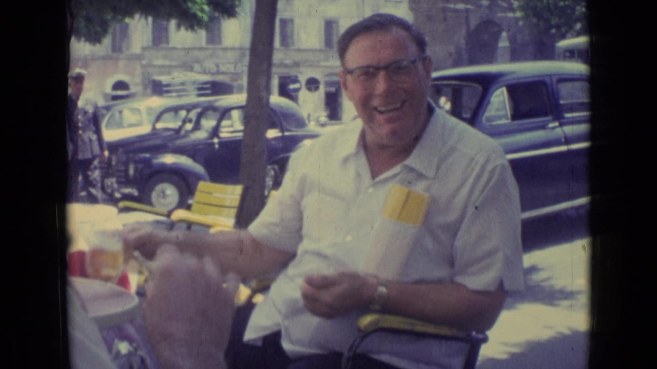A smiling man in glasses sits at an outdoor cafe table in Vatican City, framed by yellow chairs, tree shade, and mid-century parked cars.