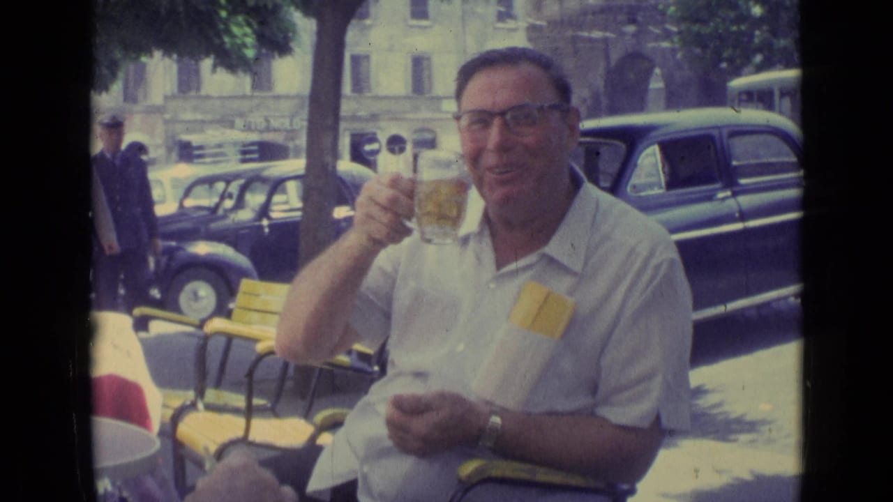 In Vatican City in 1962, a man in glasses smiles with a drink at an outdoor cafe, with yellow chairs and vintage cars behind him.