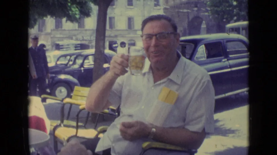 In Vatican City in 1962, a man in glasses smiles with a drink at an outdoor cafe, with yellow chairs and vintage cars behind him.