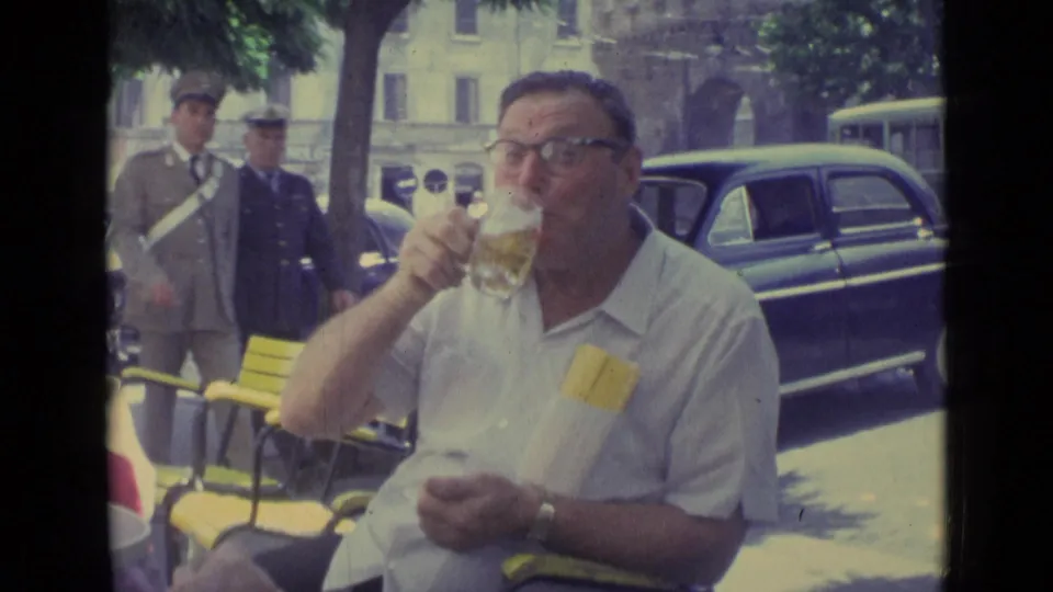 The man sips from his glass at the cafe while uniformed men pass and mid-century cars line the Vatican City street.