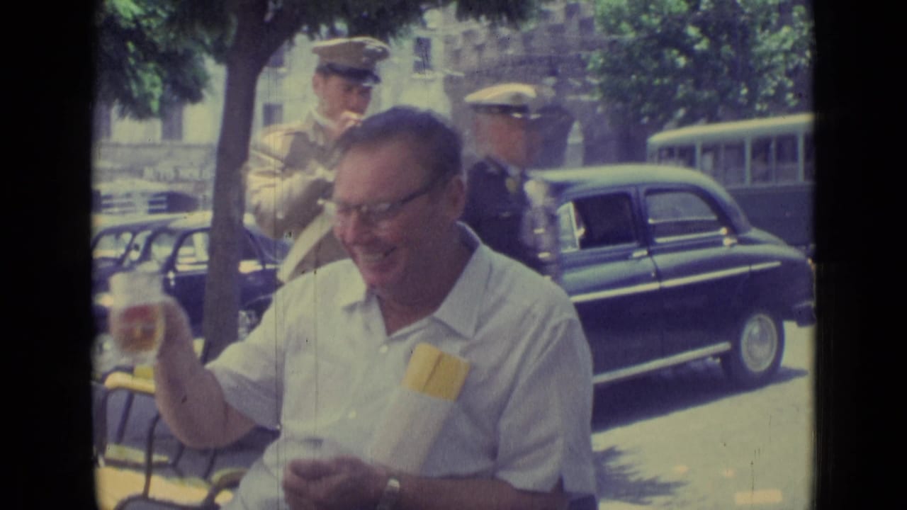 At the outdoor cafe, the man smiles while lowering his glass as two uniformed men pass behind him near parked cars.