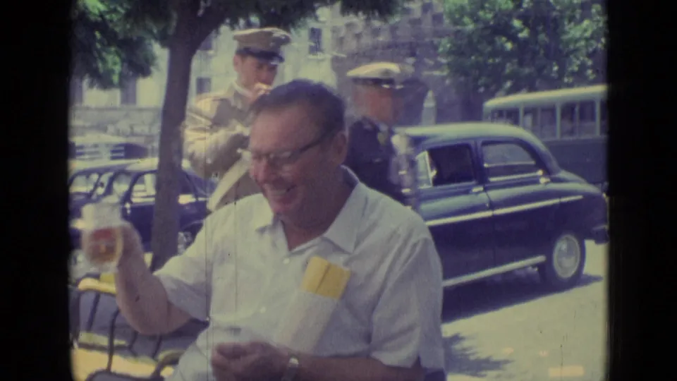 At the outdoor cafe, the man smiles while lowering his glass as two uniformed men pass behind him near parked cars.