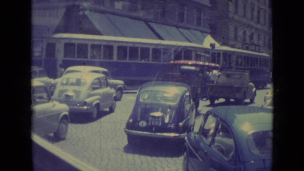 A 1962 Vatican City street view shows vintage cars and a truck on cobblestones as a blue and white tram travels along the road.