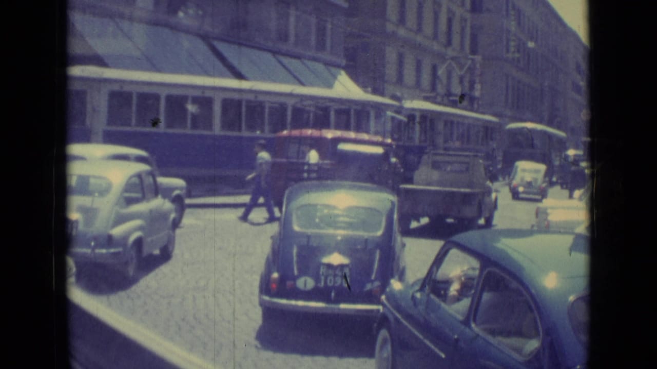 Trolley and compact cars move along a cobblestone city street.