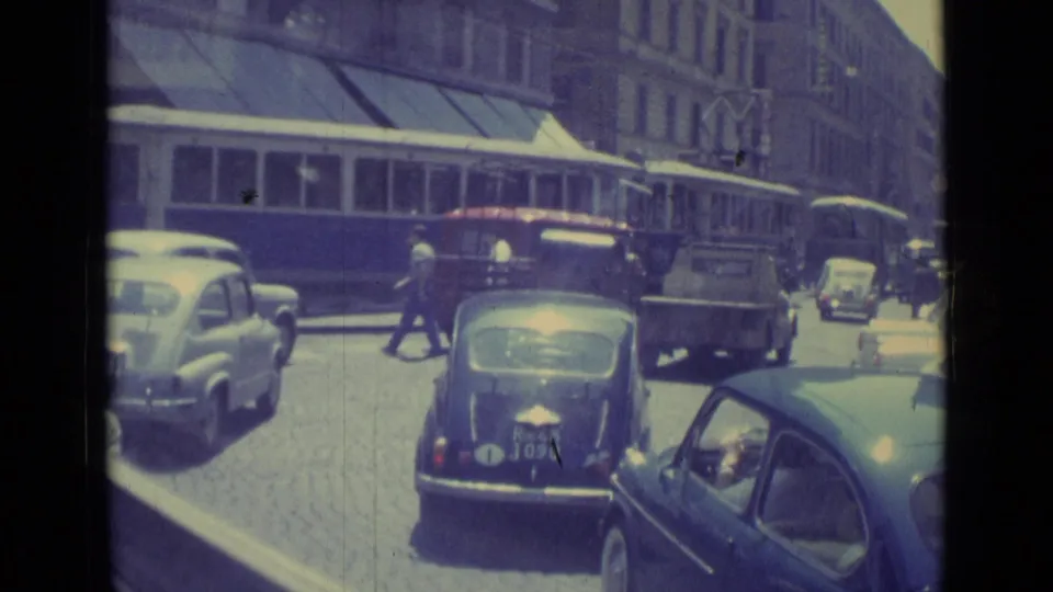 1962 Vatican City street view with a trolley, compact cars, pedestrians, awnings, and multi-story buildings in bright daylight.