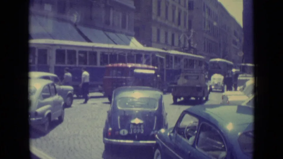 1962 Vatican City traffic scene with a trolley, parked and moving compact cars, pedestrians, and storefront awnings.