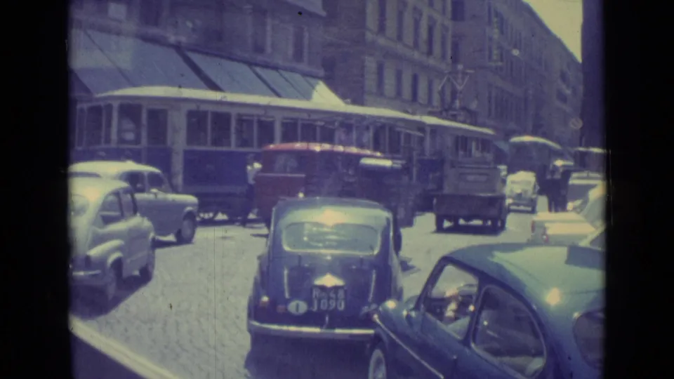 1962 Vatican City home-movie frame showing cars, a trolley, pedestrians, cobblestones, and shaded storefronts.