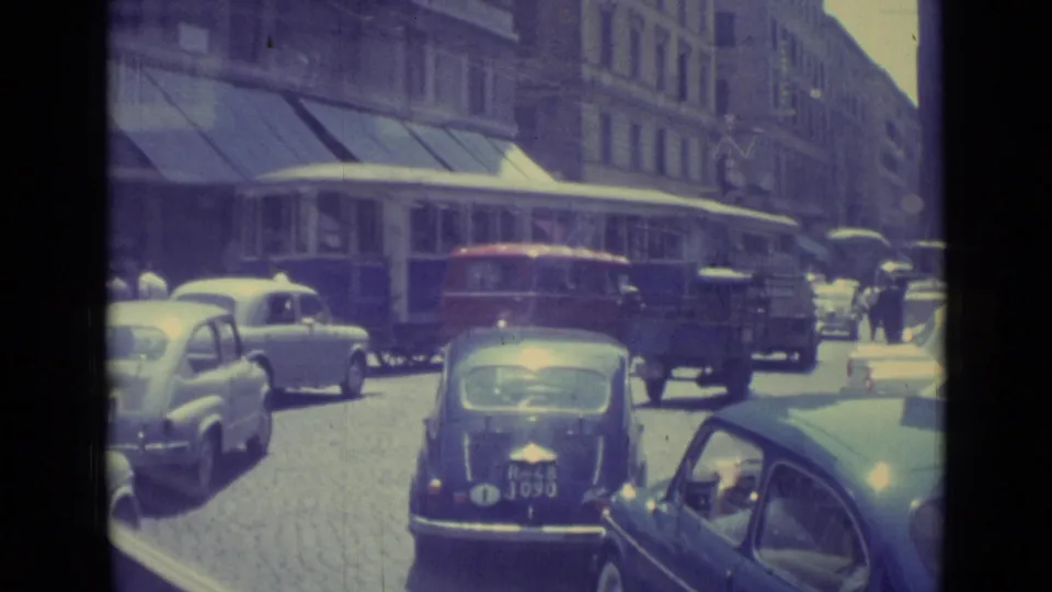 1962 Vatican City street traffic with a trolley, classic compact cars, pedestrians, cobblestones, and urban facades.
