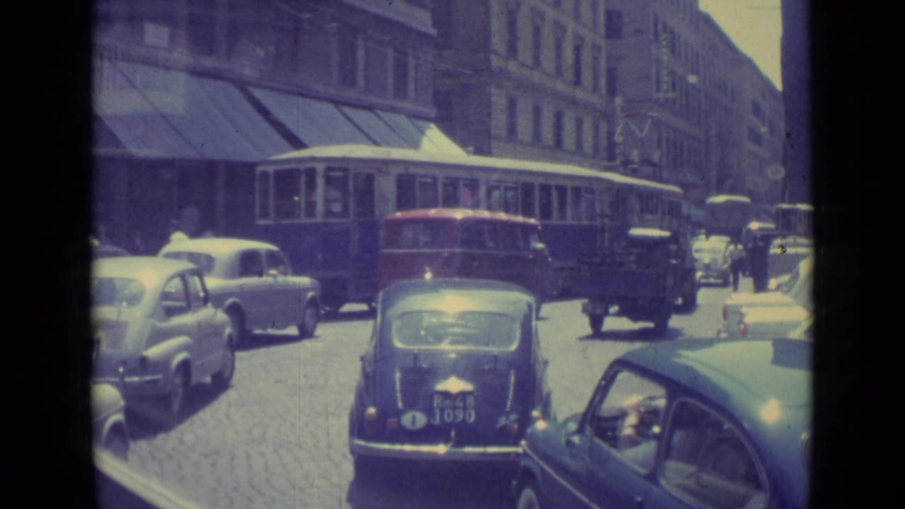 Mid-century cars and a trolley or bus move along a cobblestone city street.