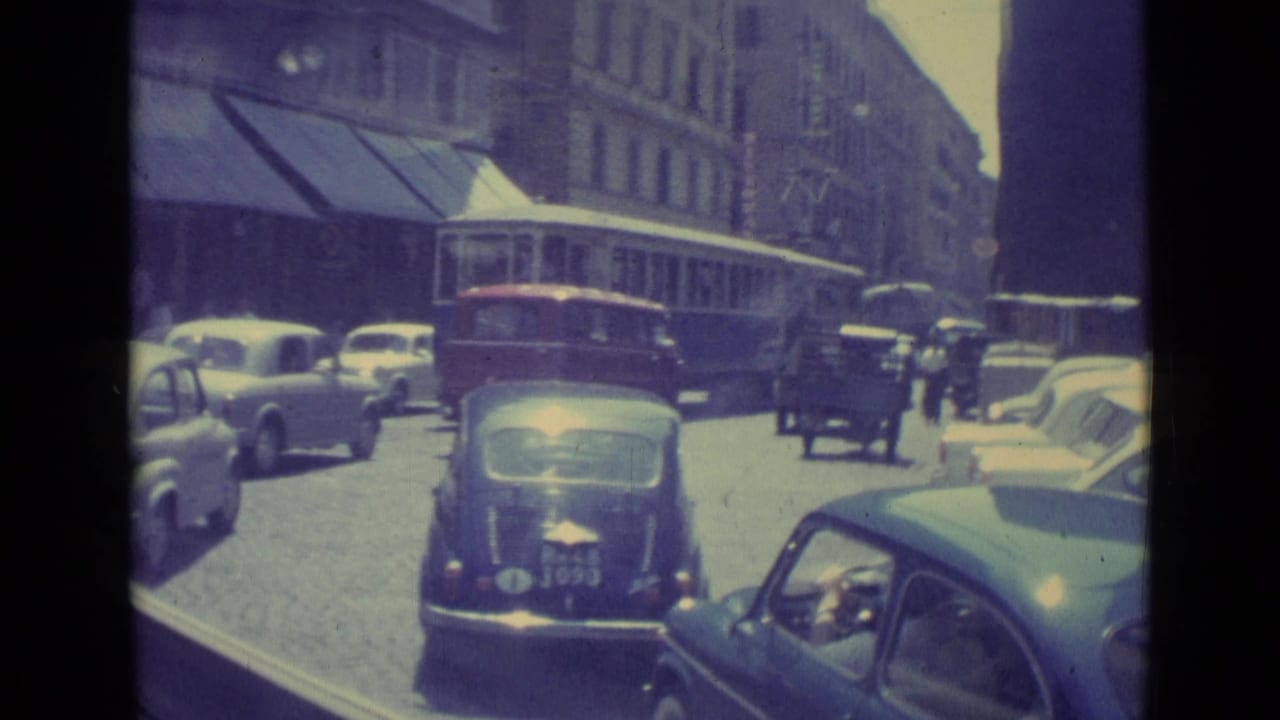 1962 Vatican City traffic scene with several compact cars, pedestrians, and a trolley or bus beside awning-covered storefronts.