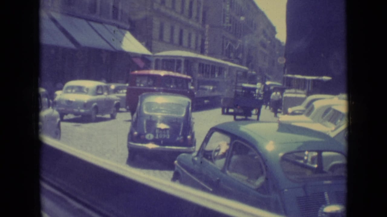 1962 Vatican City street view with a dark compact car in the lane, a trolley or bus ahead, and parked cars along the curb.