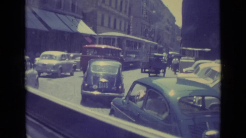 1962 Vatican City street view with a dark compact car in the lane, a trolley or bus ahead, and parked cars along the curb.