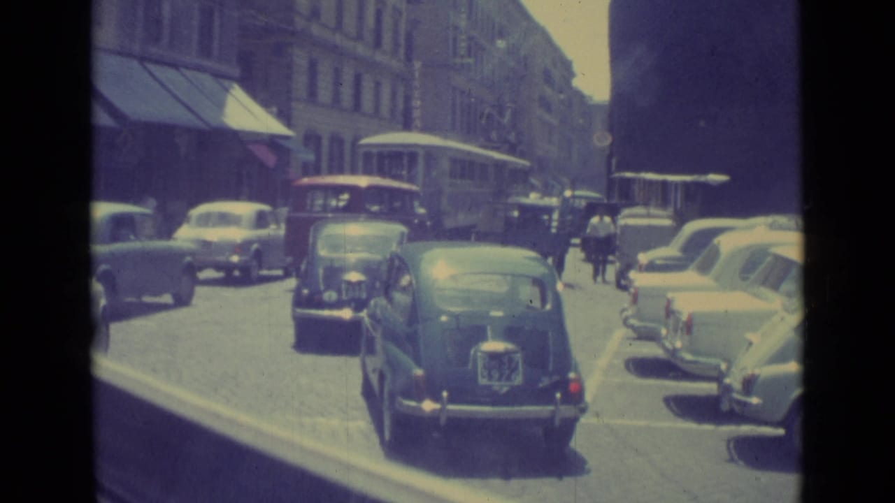 1962 Vatican City traffic with compact cars in the foreground, parked vehicles at the curb, and pedestrians near a trolley or bus.