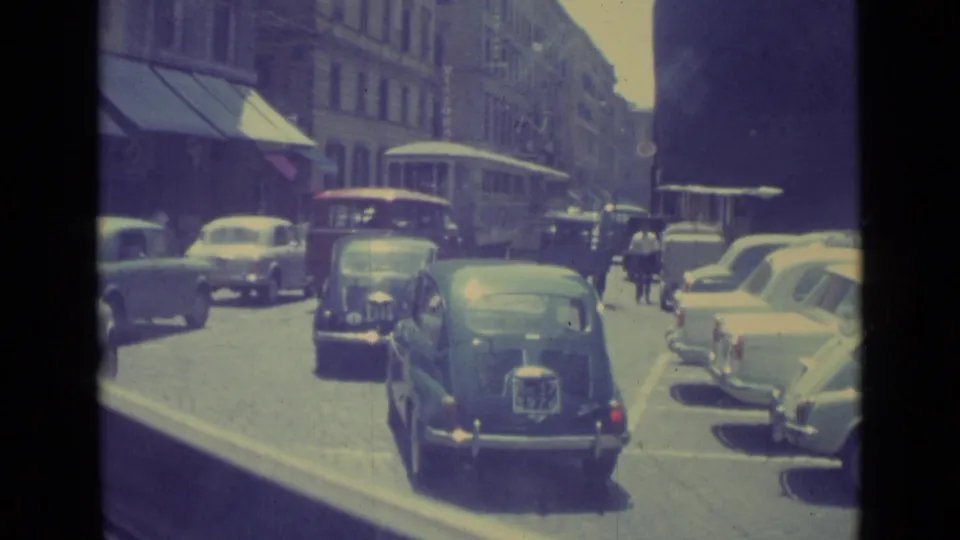 1962 Vatican City traffic with compact cars in the foreground, parked vehicles at the curb, and pedestrians near a trolley or bus.